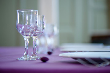 Clean table setting for fine dining with striking, elegant purple table cloth and focus on two crystal glasses for water and wine, white china and cutlery, perspective with shallow depth of field