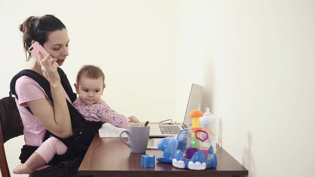 Busy Mother With Baby In Sling Carrier at home office working on laptop talking phone feeding multitasking