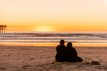 Naklejka na ściany i meble Young couple sitting on the beach watching golden sunset.