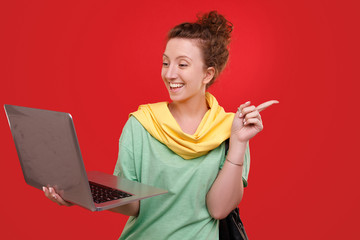 Cheerful young stylish beautiful woman holding a laptop in her hands and pointing to an empty place posing on a red background. Advertising space