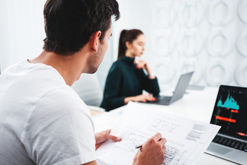 Team of male and female architect designer in office working on notebook and building blueprint. Man and woman engineers looking at drawing and computer while sits by the table in loft office