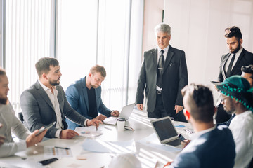 Handsome Senior grey-haired caucasian Executive leading the meeting of international partners in boardroom, having positive mood and introducing the new board member to his colleagues