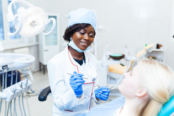 Female black dentist in dental office talking with female patient and preparing for treatment.