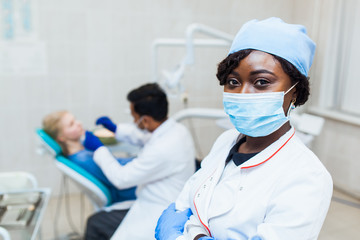 African dentist in mask looks into camera against the background of a doctor treats teeth