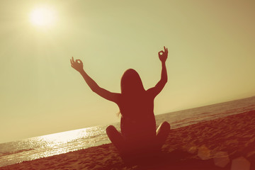 Girl meditates in the lotus position on the beach at sunset, hands up, bright beam, the horizon is twisted