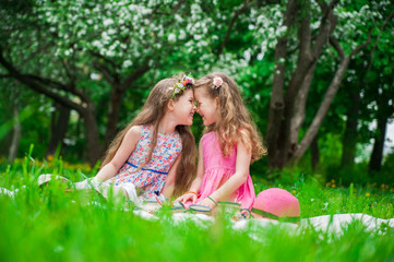 Two beautiful girls sitting on a blanket on the grass in blooming apple blossom garden.