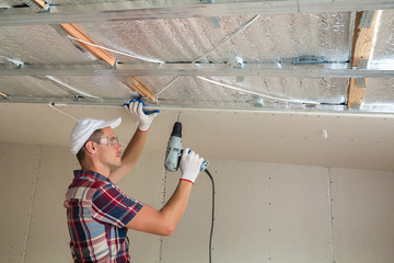 Young man in goggles fixing drywall suspended ceiling to metal frame using electrical screwdriver...