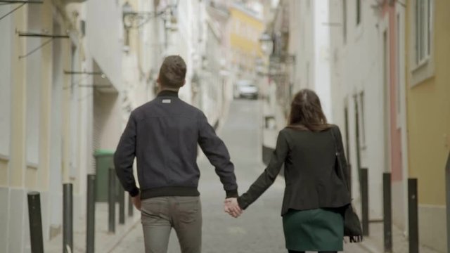 Rear view of young couple holding hands together while walking on street. Slow motion shot of young people walking at unknown city and looking around. Travel concept