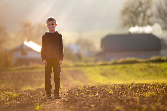 Young blond serious tired child boy standing alone on field after harvest on late summer or autumn sunny day on distant foggy blurred blue panorama of small houses among green trees background.