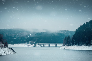 View of a mountain dam lake with a bridge in the background in a winter landscape and clouds and mist. Harz Mountains National Park in Germany