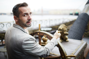 Confident businessman in suit looking at you while steering sailing wheel on large ship in urban...