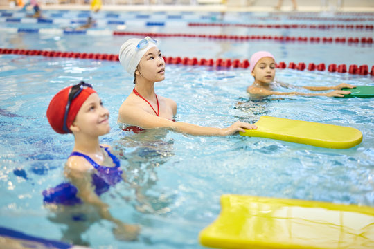 Group of girls in swimwear standing in swimming pool with kickboards and listening to their trainer during swimming lesson