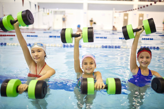 Portrait of happy little girls standing in the water and doing aerobics with dumbbells in the swimming pool