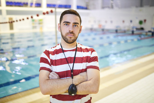 Portrait of serious young swim instructor with stopwatch on his neck standing with arms crossed in indoor swimming pool