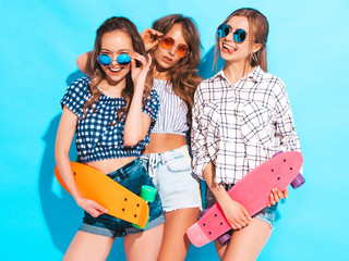 Three young stylish sexy smiling beautiful girls with colorful penny skateboards. Women in summer hipster checkered shirt clothes posing near blue wall in sunglasses. Positive models having fun
