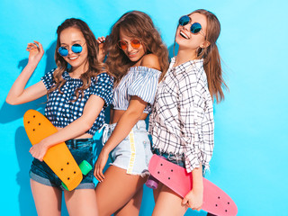 Three young stylish sexy smiling beautiful girls with colorful penny skateboards. Women in summer hipster checkered shirt clothes posing near blue wall in sunglasses. Positive models having fun
