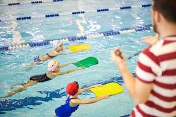 Group of little girls learning to swim with boards while swimming instructor training them