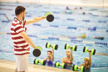 Young swim instructor standing with dumbbells and training little girls in the swimming pool