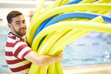 Portrait of young smiling swim instructor carrying swim noodles for his swimming class