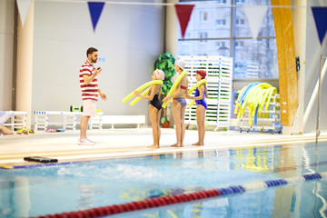 Group of girls standing with swim noodles and listening to their instructor before going to swim in the pool