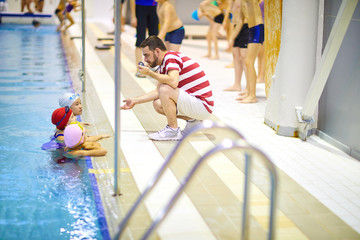 Young swim instructor sitting near the edge of the pool and has a swimming lesson with group of...