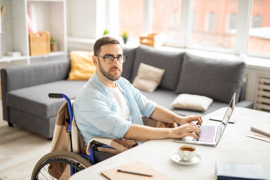 Young casual businessman in wheelchair looking at you while sitting by desk in front of laptop and networking
