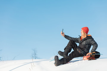 cheerful blond man in winter snowboard clothing sitting and taking a picture of himself on top of...