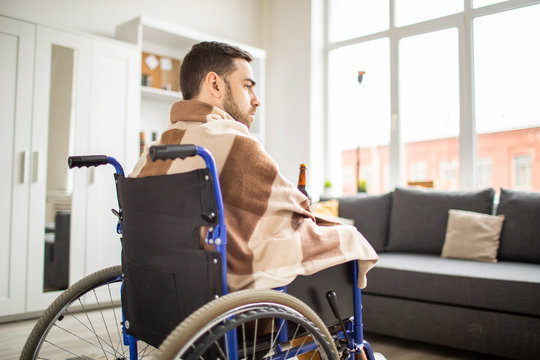 Young unhappy handicapped man in wheelchair wrapped in plaid having alcohol while spending time at home