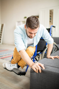 Casual disable man on wheelchair trying hard to remove on couch while holding tight by edge of sofa