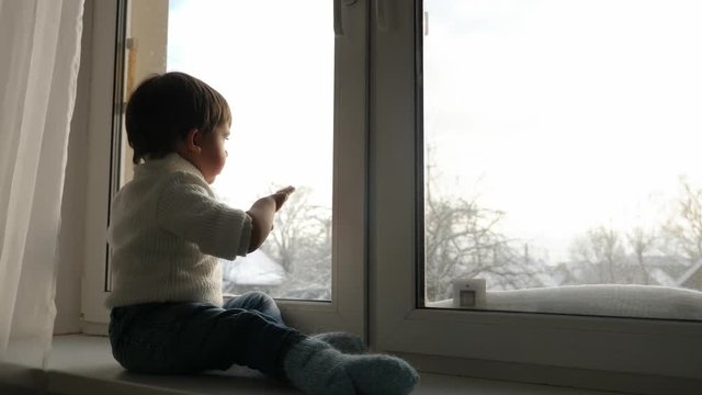 boy sits at a large window in the winter and waving