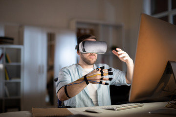Young contemporary businessman with sensors on fingers and vr goggle on head sitting in front of computer monitor