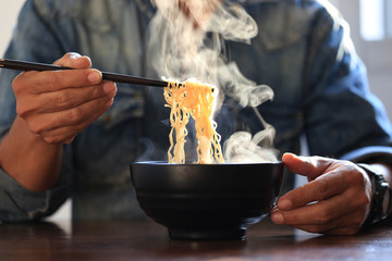 Man hand holding chopsticks of instant noodles in wood bowl with smoke rising in the home, Sodium...