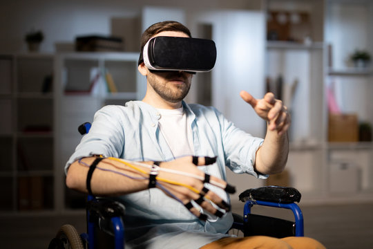 Young man with vr headset and sensors on fingers sitting in wheelchair and traveling in another reality