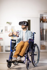 Young disable man with sensors on fingers and vr headset sitting in wheelchair while looking at curious stuff on virtual display