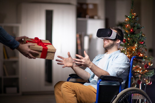 Cheerful disable man with vr goggle sitting in wheelchair and stretching arms to take giftbox with xmas present