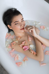 Obraz na płótnie Canvas Slim nude brunette woman enjoying bath with flower petals with closed eyes, has aromatherapy, coveres her naked body with crossed arms.