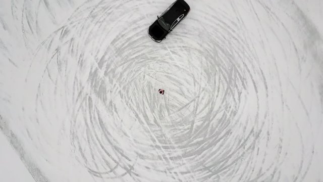 Top view rotation shot sliding and drifting black sport car moving on circle person standing in center. High angle winter race competition surrounded by white snow and ice aerial shot