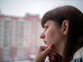 Young pretty brunette woman with long hair looks thoughtfully while standing at the window close-up, against the background of houses