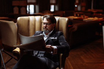 Adult man in his fifties, successful entrepreneur in spectacles and suit reading newspaper while sitting in luxury genuine leather arm-chair in dark room with lighted lamps