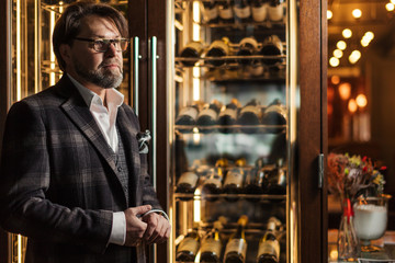 Portrait of a handsome mature male sommelier in tailored suit posing in front of the shelves with...