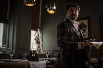 Fashionably dressed mid-aged bearded caucasian male successful clothing designer reading latest news in newspaper while sitting in restaurant.