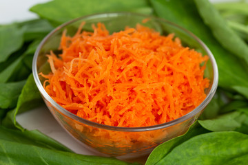 Grated carrots in a bowl on a white wooden table near the leaves of greens.