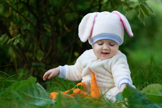 Small girl in bunny suit eating on grass in park and playing with toy carrots.
