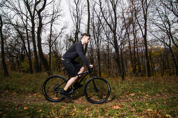 Fototapeta premium Handsome male biker in professional sportswear while riding bike down park alley on autumn day. Sportsman training thinking about future win in contest.