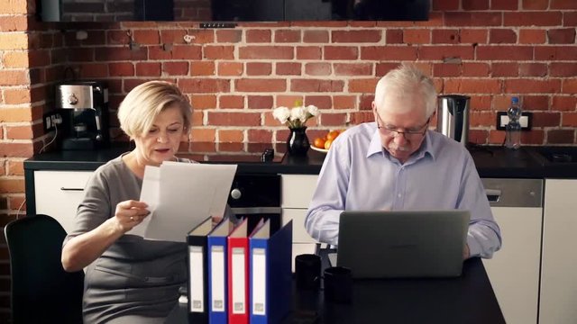 Senior couple comparing data on laptop and documents in the kitchen
 - Powered by Adobe