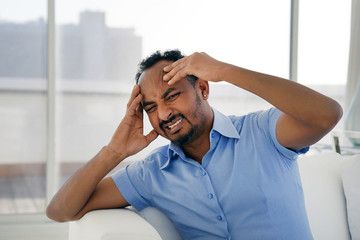 African man suffering from headache migraine pain at home on sofa. Health problem, stress and depression. Male holds head with hand. Concept of health.