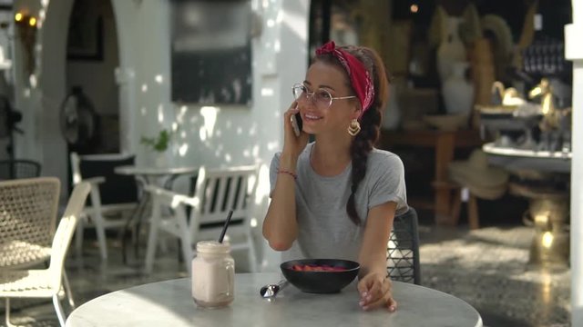 Beautiful young brunette in round glasses with plait, wearing red do-rag sitting in sunny day in outdoor cafe speaking by her phone