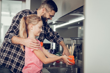 Long-haired pretty girl in a pink shirt and her father washing vegetables in the kitchen