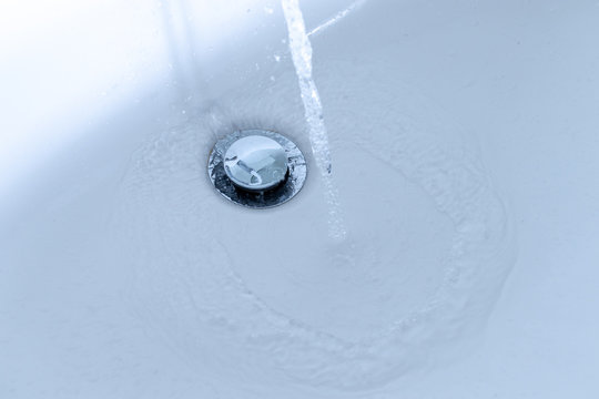 Water jet flowing in the white sink beside drain in bathroom