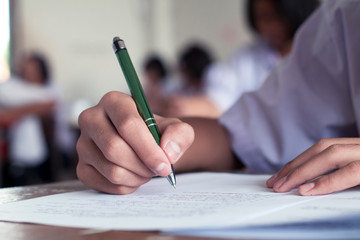 Close-up to student holding pen and writing final exam in examination room or study in classroom.
